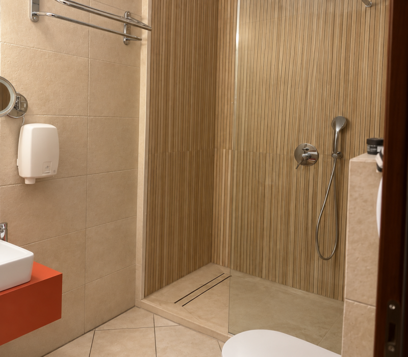 Thalassa renovated bathroom with glass shower, beige wall cladding, red vanity and white toilet. / Ανακαινισμένο μπάνιο Θάλασσας με γυάλινη ντουζιέρα, μπεζ επένδυση και κόκκινο νιπτήρα.